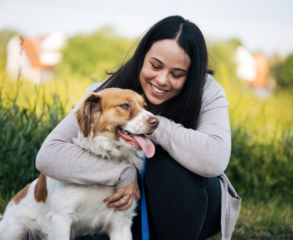 A person hugging a dog outdoors, with a blurred background of greenery and buildings