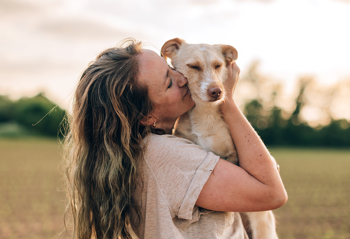 close up portrait of a happy woman hugging and kissing her dog Woman with long hair lovingly kisses a tan dog outdoors at sunset