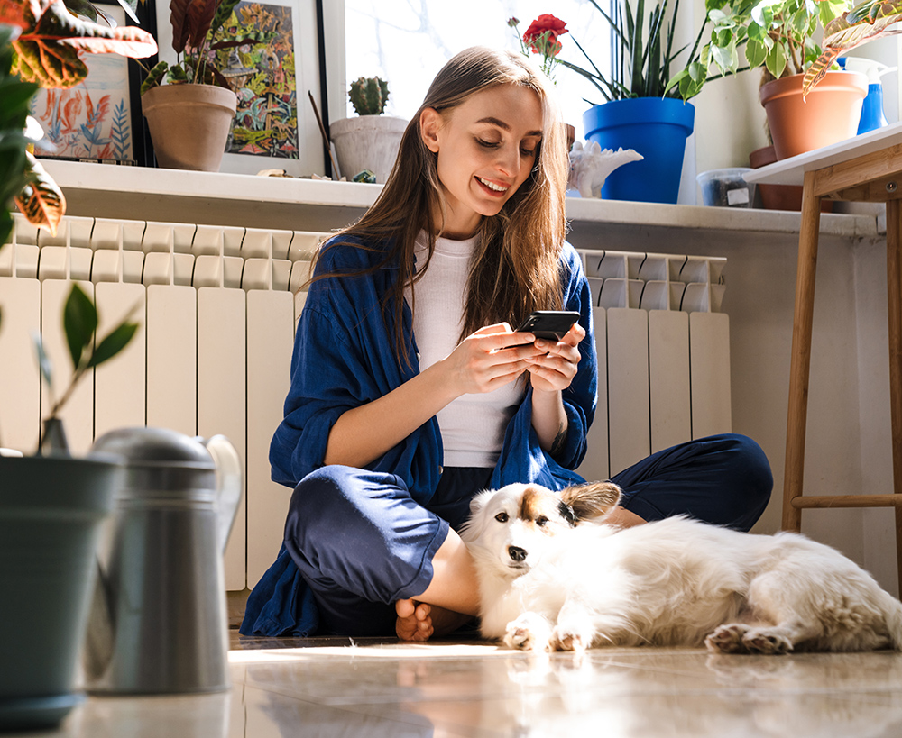 A woman sitting cross-legged on the floor, smiling at her phone, with a fluffy dog resting beside her.